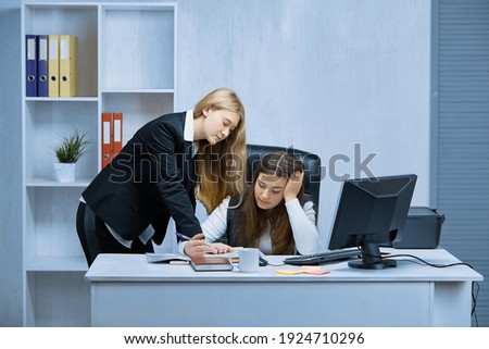 Two girls at a white desk in the office, consulting while reviewing a shared project. In the background is a rack of documents.
