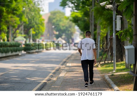 Back view of a young man wearing a white t-shirt and long black trousers walking on the footpath. Royalty-Free Stock Photo #1924761179