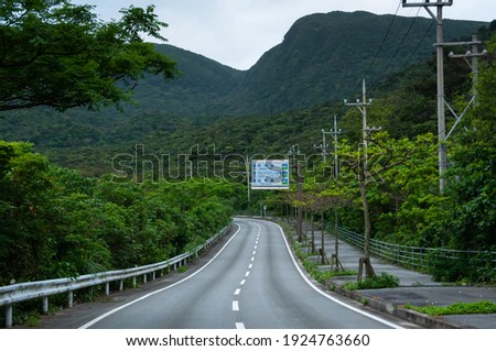 Tropical island road, lush vegetation around, road sign, beautiful green mountains in the background. Iriomote Island.