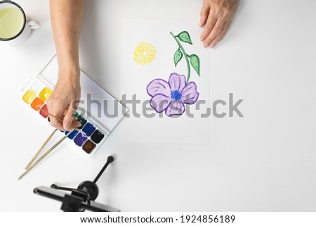 Female elderly hands paint a flower with watercolors indoors, top view. Smartphone, paints, brushes on the table. Online drawing course, retirement hobby concept