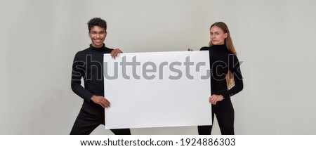 Young african american man and caucasian girl holding blank poster and smiling at camera while posing on light background, panorama. Discount and sale concept