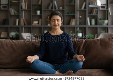 Calm millennial Indian woman sit on sofa in living room practice yoga with mudra hands. Young mixed race ethnicity female meditate relieve negative emotions relax at home. Stress free, peace concept. Royalty-Free Stock Photo #1924897085
