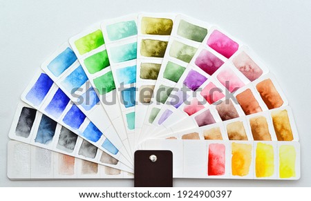 The color palette is a semicircle made of white watercolor paper with painted rectangles. Pantone fan lies on a white background. Various colors red, blue, green, yellow, metallic shades.