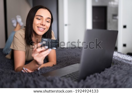 A smiling woman on her bed with laptop in front of her and credit card in hand, with crossed feet.