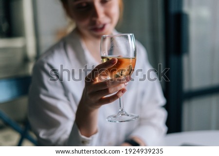 Close-up blurry photo of happy redhead young woman holding glass with champagne near face, looking away. Portrait of pretty female with glass of wine.