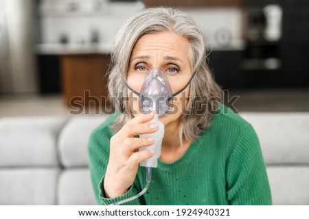Elderly woman grey-haired, has a lot of wrinkles, feels bad, holding oxygen mask, lacking of oxygen, sensitive to the polluted air, sitting at home on a self-isolation, looks sickly, doing inhalation Royalty-Free Stock Photo #1924940321