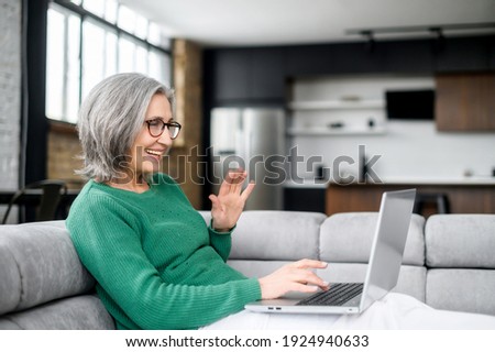 Video call concept, lovely elderly woman with nice wrinkles, narrow lips, smiling, chatting with family, using a laptop for video connection, remote meeting, say hi, glad to see, lying on the couch Royalty-Free Stock Photo #1924940633