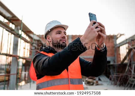 Civil engineer or architect with hardhat and orange construction vest on construction site taking photos of the progress with the smartphone camera for the report.