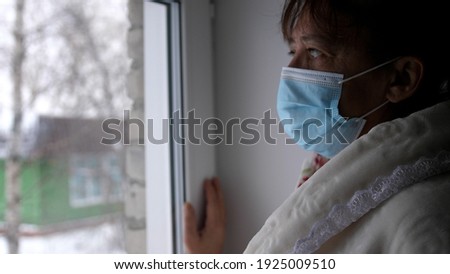 Old despair woman in protective medical mask looking sadly out window, sitting at home in quarantine. Self isolation in coronavirus pandemic. prevention of covid-19