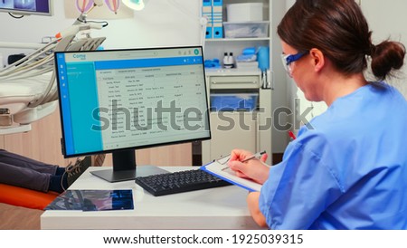 Orthodontist nurse looking at list with appointments in computer and taking notes on clipboard while dentistry doctor is working with patient background examining teeth problems..