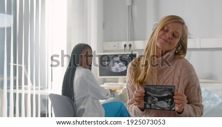 Smiling pregnant woman holding ultrasound baby image sitting in gynecologist office. Happy caucasian female holding sonogram picture with african doctor sitting on background