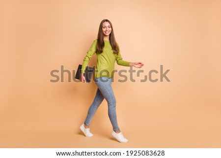 Photo of girl walk hold computer device wear green pullover jeans sneakers isolated beige color background