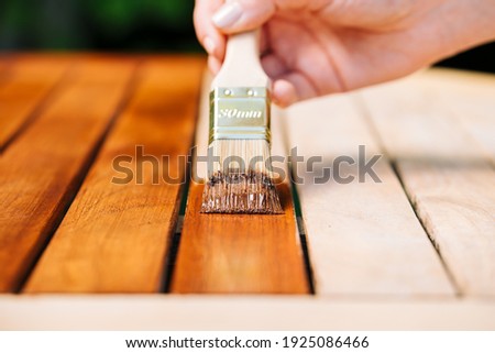 hand holding a brush applying varnish paint on a wooden garden table - painting and caring for wood with oil Royalty-Free Stock Photo #1925086466