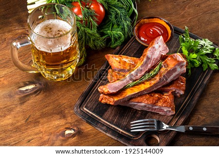  Smoked pork ribs with ketchup on a cutting board and a glass of light beer on the wooden background