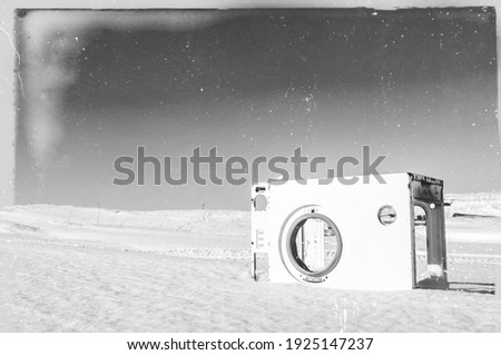 Old, damaged vintage retro style tundra landscape black and white photo. Old washing machine covered with snow in the front. Lots of age marks, letters, scratches placed on the film tape surface. 