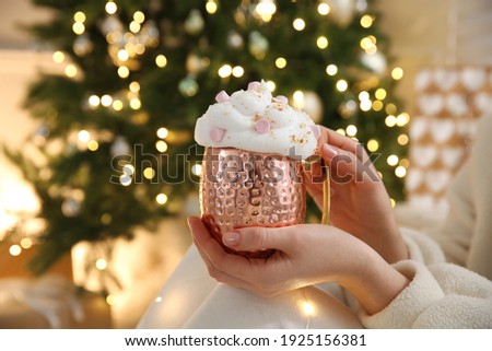 Woman holding cup of delicious drink with whipped cream and marshmallows near Christmas tree indoors, closeup