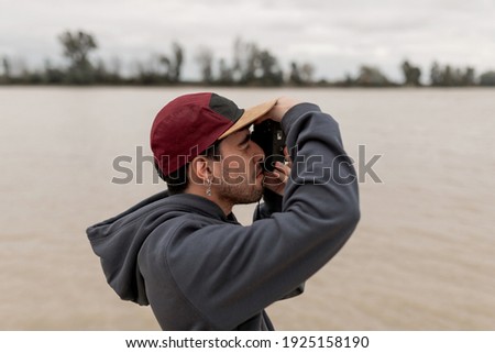 Young man wearing a grey sweater and red cap is taking photos in a pier next to a river