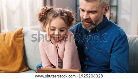 Close up of caring loving Caucasian father with little daughter spending time together at home using tablet. Dad teaching small child girl to use gadget device, watching cartoons, family concept