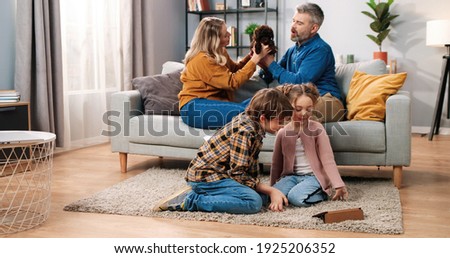 Joyful Caucasian family spending time at home. Happy kids little boy and girl sitting on floor watching videos or cartoons on tablet while parents playing with pet puppy dog on sofa, leisure concept