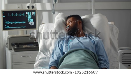 Seriously ill african man lying on bed in hospital with monitors showing his vital signs. Portrait of sad afro-amerian male patient resting in intensive care unit
