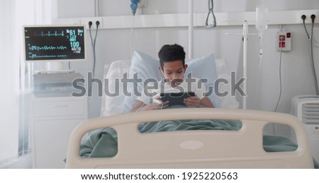 African kid patient using digital tablet on bed in hospital. Portrait of preteen afro-american boy lying in bed and playing game on tablet pc in hospital ward
