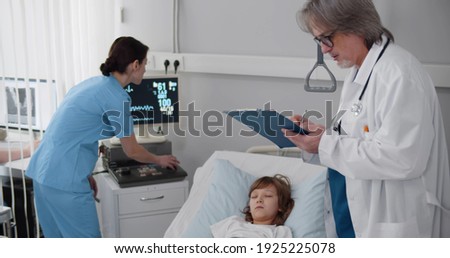 Senior doctor and nurse checking sick child patient sleeping in hospital bed. Medical team checking vital signs of ill preteen boy lying unconscious in hospital ward