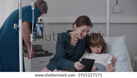 Preteen boy resting in bed at children hospital with mother sitting beside watching cartoons on tablet together while doctor checking monitor in hospital ward