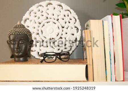reading glasses and black buddha head on a book lying on the library shelf on grey background and white mandala and green plant