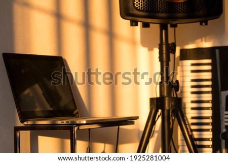 A laptop on a metal desk with music equipment like a PA speaker on a stand and a piano keyboard blurred in the background in a studio room during sunset. Royalty-Free Stock Photo #1925281004