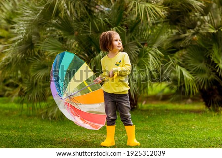 beautiful little girl.in a yellow sweater, jeans,and yellow boots, he stands with a colored umbrella against a background of green palm trees