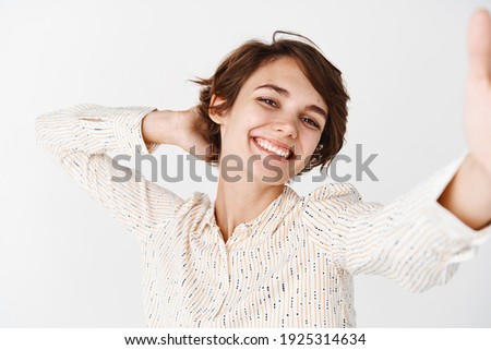 Candid happy woman posing for selfie, holding smartphone and taking pic of herself with cheerful smile, standing carefree on white background.