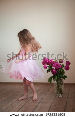 A cute little girl with long hair in a beautiful pink dress is dancing near a vase with roses that have faded a little