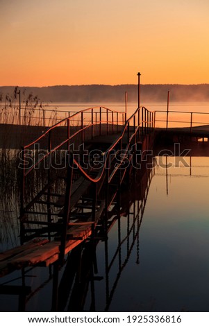 A magical beautiful early morning on the lake in spring. An old wooden abandoned pier. Sunrise over the water with fog. Beautiful orange sky. Marina for boats. Romantic place.