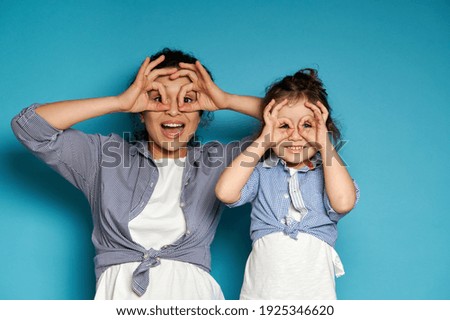 Happy woman and her cute daughter looking at camera through their fingers in ok gesture, imitating binoculars. Copy space, blue background. Royalty-Free Stock Photo #1925346620