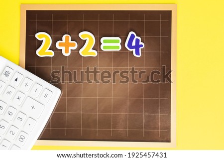 Next to a white calculator on a yellow background, there is a school magnetic board with a correct example of colored numbers on it. The concept of education.