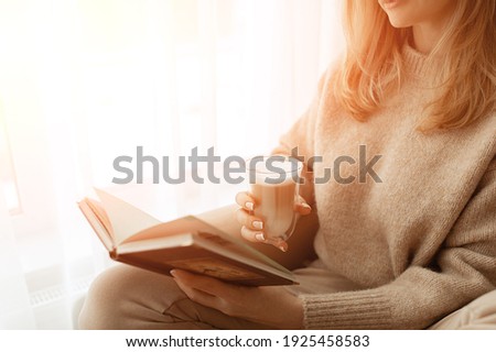 Young beautiful woman near window beige knitted sweater read book, wish list, daily planner, notepad, goals. Relax concept. Hold cappuccino glass of coffee with white foam. Sun's rays shine into room Royalty-Free Stock Photo #1925458583