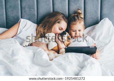 Baby girl and her mother with dog lying on the bed and watching on tablet. Family spending time together. 