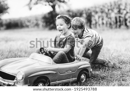 Two happy children playing with big old toy car in summer garden, outdoors. Boy driving car with little girl inside. Laughing and smiling kids. Old picture in black and white