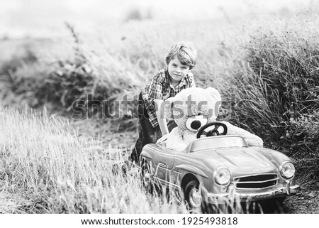 Little preschool kid boy driving big toy car and having fun with playing with his plush toy bear, outdoors. Child enjoying warm summer day in nature landscape. Old picture in black and white.