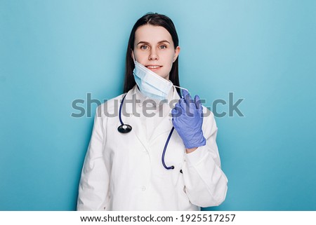 Professional female doctor in latex protective gloves removing face mask, wears white uniform and stethoscope, isolated on blue background. Covid 19, healthcare workers and preventing virus concept