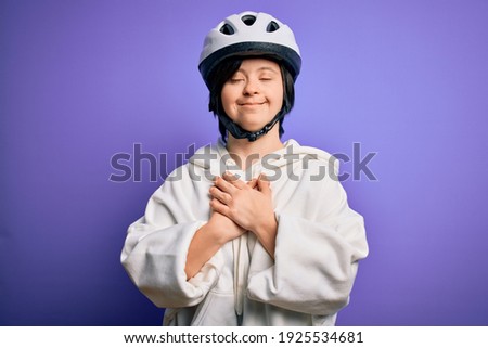 Young down syndrome cyclist woman wearing security bike helmet over purple background smiling with hands on chest with closed eyes and grateful gesture on face. Health concept.