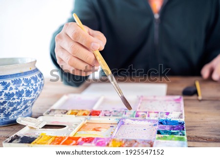 Man artist painting with paint brush and watercolor palette on paper in front of laptop, closeup. Selective focus.