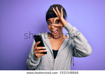 African american sporty woman doing sport listening to music using smartphone and earphones with happy face smiling doing ok sign with hand on eye looking through fingers