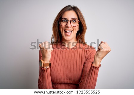Young beautiful brunette woman wearing casual sweater and glasses over white background celebrating surprised and amazed for success with arms raised and open eyes. Winner concept.