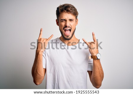 Young handsome man with beard wearing casual t-shirt standing over white background shouting with crazy expression doing rock symbol with hands up. Music star. Heavy concept.