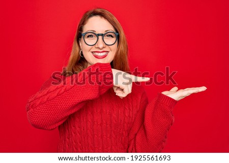 Young beautiful redhead woman wearing casual sweater and glasses over red background amazed and smiling to the camera while presenting with hand and pointing with finger.