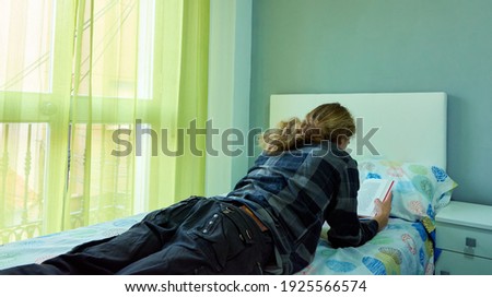 An unrecognizable male student with long hair lying in his bed and reading a book in a bedroom