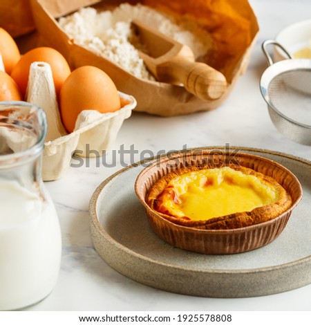 Egg tart, traditional Portuguese dessert, pastel de nata in a gray plate on a marble background . Portuguese puff and ingredients for cooking (flour, butter, eggs, milk). square picture