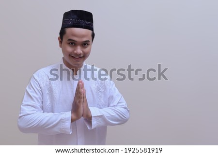 Portrait of religious Asian man in koko shirt or white muslim shirt and black cap, showing apologize and welcome hand gesture. Isolated image over gray background