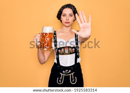Beautiful german woman with blue eyes wearing traditional octoberfest dress drinking jar of beer with open hand doing stop sign with serious and confident expression, defense gesture